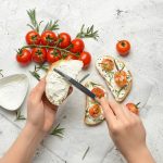 Hands spread cream cheese on sliced bread as cherry tomatoes, rosemary, and sesame seeds are arranged on top for crostini on parchment.