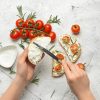 Hands spread cream cheese on sliced bread as cherry tomatoes, rosemary, and sesame seeds are arranged on top for crostini on parchment.