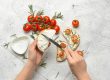 Hands spread cream cheese on sliced bread as cherry tomatoes, rosemary, and sesame seeds are arranged on top for crostini on parchment.