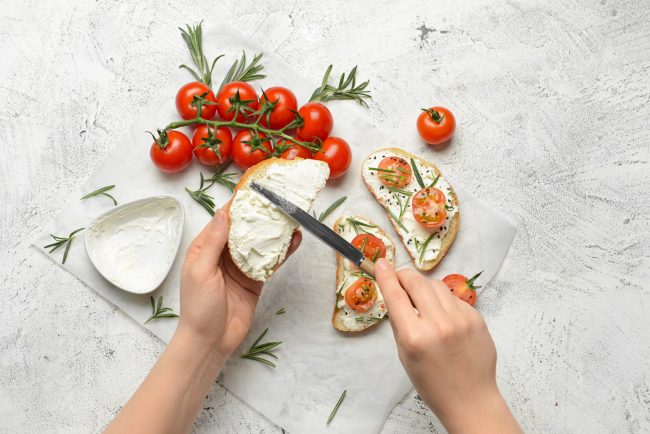 Hands spread cream cheese on sliced bread as cherry tomatoes, rosemary, and sesame seeds are arranged on top for crostini on parchment.